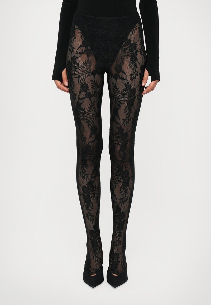 Meia-calça preta e transparente com padrão de renda floral; cintura alta; textura suave; combinada com sapatos de salto alto pretos.