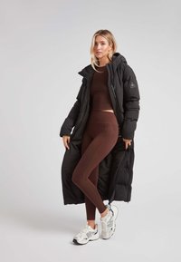 Abrigo de plumas negro con capucha, llevado sobre una camiseta cortada marrón y leggings marrones, combinado con zapatillas blancas.