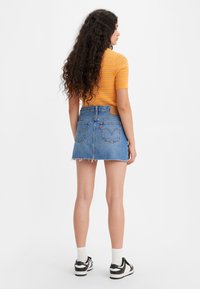 Spijkerrok met een rafelige onderkant, blauwe kleur, met twee achterzakken en een bruine leren label. Gecombineerd met een gestreept oranje topje en sneakers.