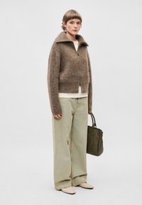 Sweater castanho felpudo com fecho, calças largas verde-claro, sapatilhas bejes de bico apontado e uma mala verde-escura. Design simples e textura suave.