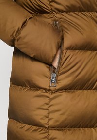 Una mano se desliza en el bolsillo con cremallera de una chaqueta de invierno acolchada marrón, mostrando el detalle en plata de la cremallera y la textura de la tela.