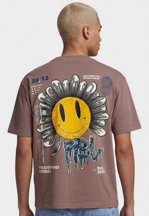 Person med kort blondt hår har en brun T-shirt på med en stor smilende blomster og streetwear-inspireret tekst og grafik på ryggen.