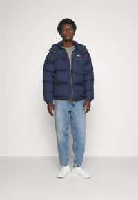 Giacca a vento blu marina con zip anteriore, cappuccio e tasca frontale in contrasto. Indossata sopra un maglione grigio e abbinata a jeans azzurri chiari e scarpe bianche.