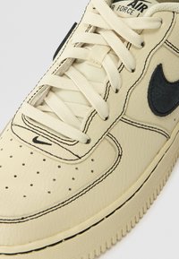 Sapatilha de couro creme com detalhes em preto, caixa do dedo perfurada, atacadores planos e costura contrastante. Apresenta o logótipo da Nike na lateral e na língua.
