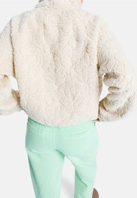 Kremowa kurtka z polaru o teksturze kremu z wysokim kołnierzem, ozdobiona kwiatowym wzorem, połączona z miętowozielonymi spodniami. Widok z tyłu ukazuje swobodny krój.