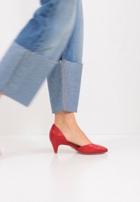 Κόκκινο δερμάτινο παπούτσι με ψηλό τακούνι, μυτερή μύτη και καμπύλο τακούνι. Φορεμένο με μπλε τζιν παντελόνι φαρδιάς γραμμής με ξεφτισμένα τελειώματα.