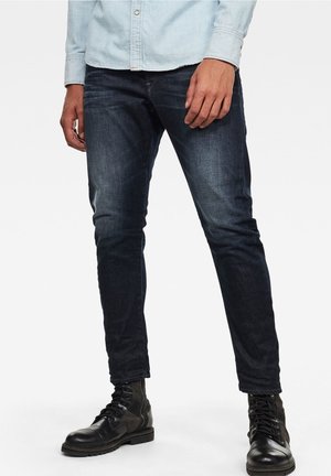 Vaqueros de mezclilla oscuros con un corte ajustado, que presentan un leve desgaste y costuras decorativas. Combinados con botas negras de cordones.