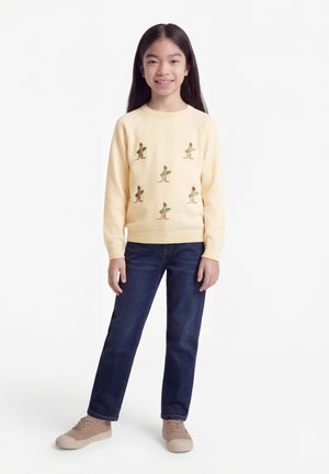 Κορίτσι που στέκεται χαμογελώντας, φοράει πουλόβερ κρέμα με σχέδια καγκουρό και φύλλων, σκούρα μπλε τζιν και μπεζ αθλητικά παπούτσια.