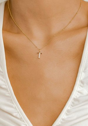 Gold necklace with a small diamond-encrusted 'T' pendant, featuring a delicate chain with round beads at intervals, set against a light fabric background.