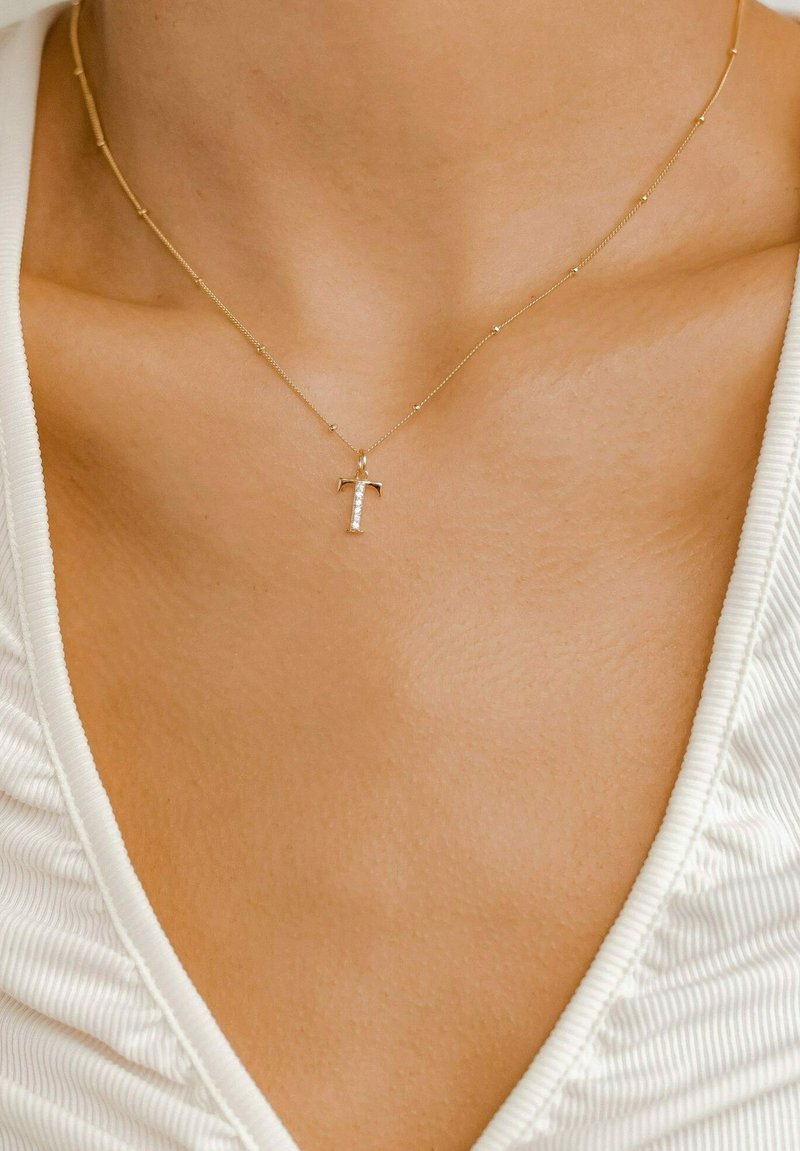 Collar de oro con un pequeño colgante en forma de 'T' engastado con diamantes, que presenta una delicada cadena con cuentas redondas a intervalos, sobre un fondo de tela clara.