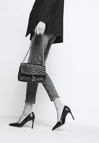 Una persona con una chaqueta negra y jeans ajustados camina con gracia, llevando un bolso acolchado negro y usando tacones altos puntiagudos.