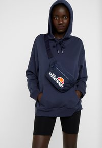 Felpa blu navy con cappuccio, coulisse e tasca frontale, abbinata a pantaloni corti neri. Una borsa a tracolla blu navy con logo bianco e dettaglio rosso.