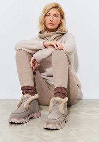 Bottines en suède gris avec bordure en fausse fourrure, semelles en caoutchouc et motif en zigzag. Associées à un pull beige à côtes et un pantalon.