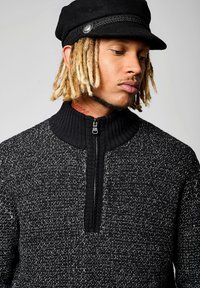 Sweat à zip noir avec un motif de tricot texturé, col montant et accents noirs. Le mannequin porte un chapeau noir avec une bande tressée.