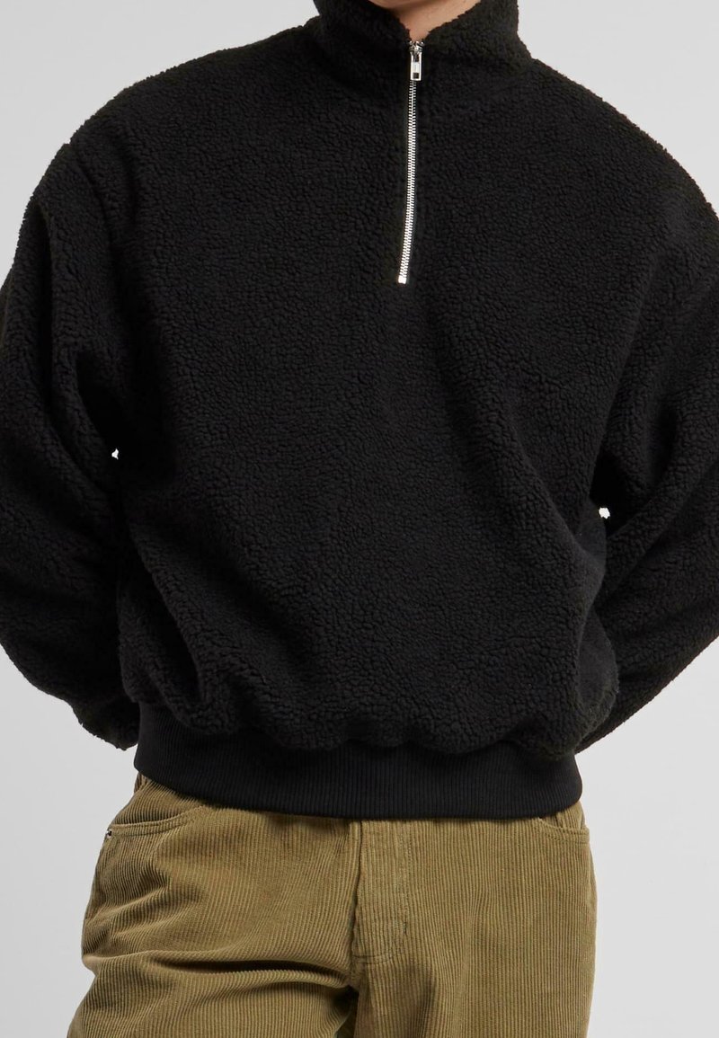 Sort fleece pullover med høj krave, halv lynlås og elastiske mansjetter og bund. Blød tekstur og afslappet pasform. Fløjl-bukser synlige forneden.