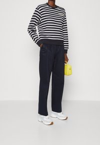 Sweatshirt bleu marine rayé avec des rayures blanches, encolure dégagée et détail de logo. Pantalons bleu marine foncé, baskets blanches avec accents bleus et sac à main jaune.