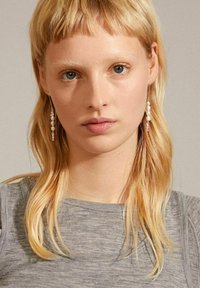 Silver dangling earrings featuring white pearls, dangling thread design, a smooth texture, and minimalist aesthetic. Model with blonde hair wearing gray top.
