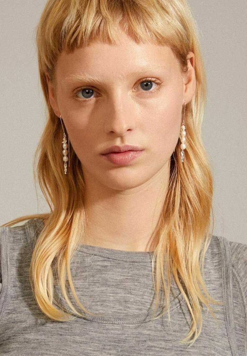 Silver dangling earrings featuring white pearls, dangling thread design, a smooth texture, and minimalist aesthetic. Model with blonde hair wearing gray top.
