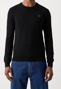 Czarny sweter z długim rękawem wykonany z gładkiej tkaniny. Posiada okrągły dekolt i małą naszywkę z logo na piersi. Połączony z niebieskimi spodniami dżinsowymi.