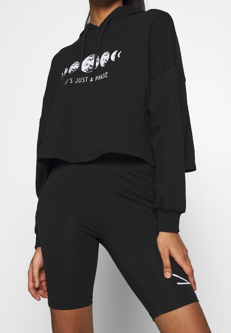 Femme portant un sweat à capuche noir court avec des phases lunaires et le texte « C’est juste une phase », associée à un short de vélo noir taille haute.