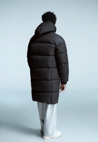 Geacă puffer neagră cu glugă și secțiuni quilted orizontale; lungime lungă, ajungând până la jumătatea gambelor, purtată peste pantaloni de trening gri și pantofi crem.