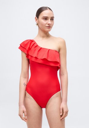 Frau mit einem roten einseitigen Badeanzug mit Rüschendetail und sternförmigen Ohrringen, stehend vor einem schlichten weißen Hintergrund.
