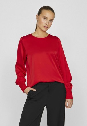 Rode satijnen blouse met lange mouwen, elastische manchetten en een ronde halslijn, gecombineerd met zwarte nauwsluitende broek. Zachte textuur, losse pasvorm.