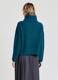 Tahrentgrüner, gerippter Strickpullover mit hohem Kragen und lockerer Passform, getragen über einem grauen, gefalteten Rock. Die Rückansicht zeigt die cropped Länge und das strukturierte Muster.
