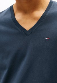 Námořnická modrá bavlněná v-krční tričko s malým logem v červené, bílé a modré na levé části hrudi. Měkká textura a neformální design.
