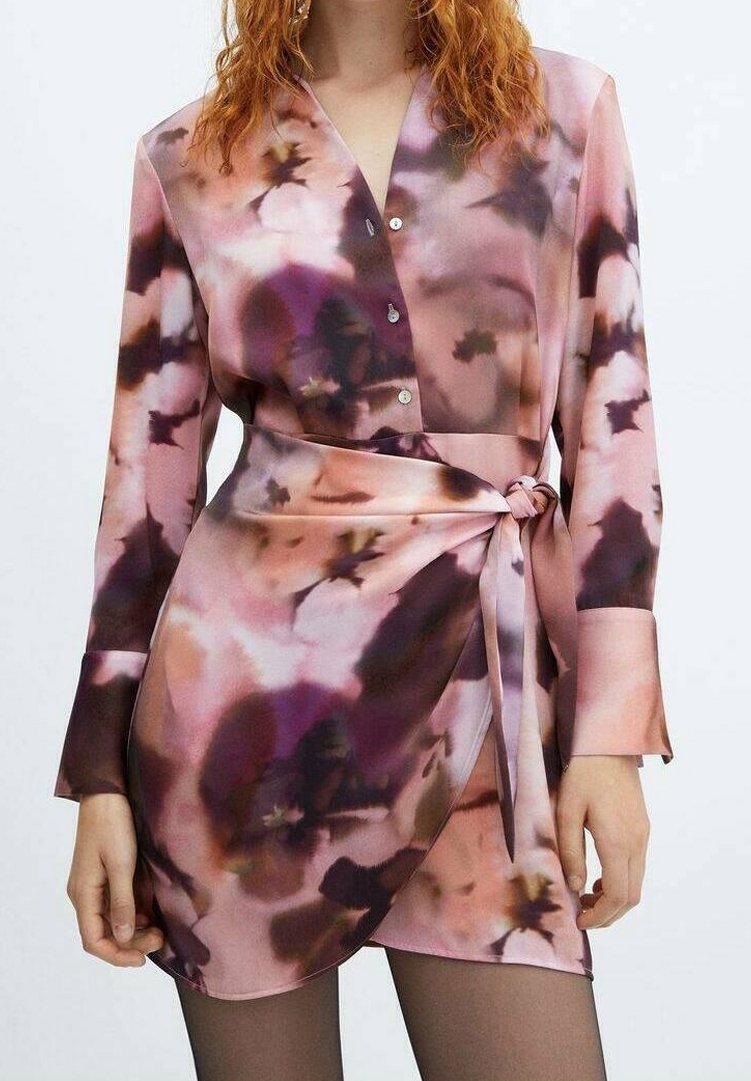 Femme portant une mini-robe à manches longues avec boutons, à motif floral abstrait rose et violet, nouée à la taille avec une ceinture assortie.