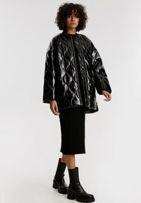 Gequilte zwarte pufferjack met een glanzende afwerking, voorrits en ribgebreide manchetten, gecombineerd met een zwarte ribgebreide midi-rok en chunky haklaarzen.