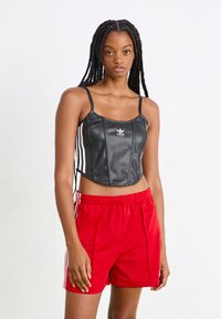 Top curto de couro sintético preto com alças finas, apresentando o logo branco da Adidas e riscas laterais. Combinado com calções de desporto vermelhos.