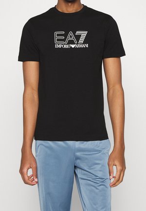 Sort t-shirt med korte ærmer med hvidt "EA7 Emporio Armani" logo, båret af en person, der står med arme afslappet, kombineret med lyseblå bukser.