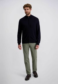 Dunkelblauer Zip-Hoodie mit einem hohen Kragen, getragen mit hellgrüner Hose und braunen Slippern. Weicher Stoff, taillierte Passform.