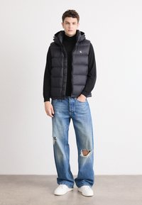 Zwarte puffer gilet met capuchon, rits aan de voorkant en logo-detail, gedragen over een zwart coltrui, gecombineerd met lichtblauwe ripped jeans en witte sneakers.