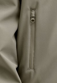 Maslinasto zelena jakna od tkanine s vertikalnim džepom na patent zatvarač s trakicom za povlačenje. Glatka tekstura i blago podstavljena konstrukcija za toplinu.