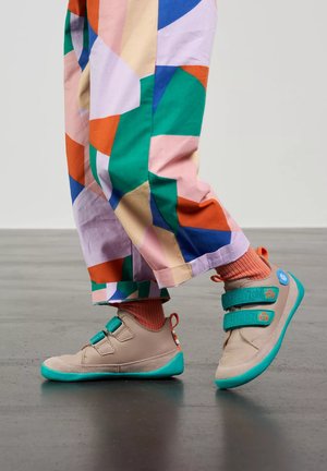 Ein Kind läuft in bunten Hosen und beige Turnschuhen mit türkisfarbenen Riemen und Sohlen, kombiniert mit orangen Socken, auf einem glatten Boden.