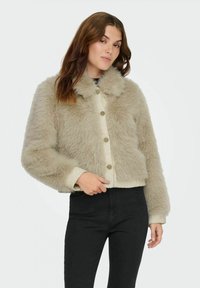 Faux fur cropped jacket in light beige with a soft texture, round collar, and large button closures. Paired with black pants.