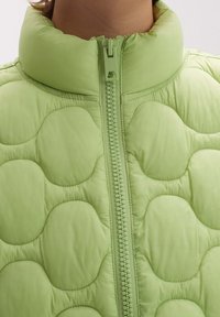 Jasnozielona kurtka puchowa z wysokim kołnierzem, z zamkiem błyskawicznym z przodu i zaokrąglonym pikowanym wzorem na całej powierzchni materiału.