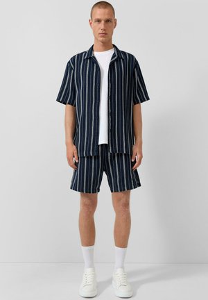 Junger Mann trägt ein kurzärmeliges Hemd mit vertikalen Streifen in Marineblau und Weiß sowie passende Shorts, ein weißes T-Shirt, weiße Socken und weiße Turnschuhe.