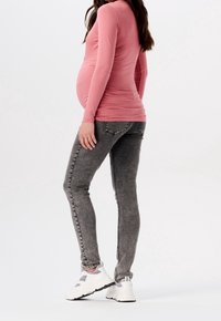 Bluzka ciążowa w jasnoróżowym kolorze, wykonana z miękkiego materiału, z długimi rękawami, zestawiona z szarymi rurkami o wyblakłej fakturze i białymi sportowymi butami.