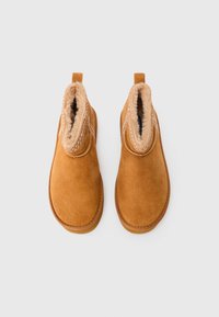 Un paio di stivali in suede beige da usare in inverno, con fodera in pelliccia beige e linguette di estrazione, mostrati dall'alto su uno sfondo bianco.