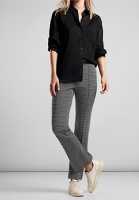 Woman wearing black buttoned shirt with rolled sleeves, gray patterned pants, and white sneakers, standing against gray background.