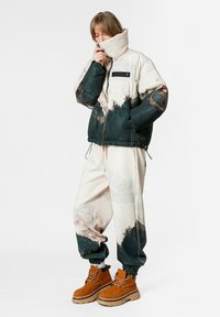 Chaqueta acolchada en beige y verde oscuro con un estampado abstracto, combinada con pantalones a juego. Botas marrones con cordones y suela gruesa.