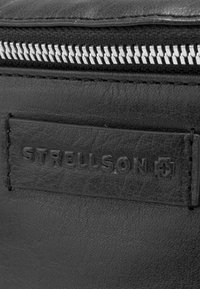 Sort lædertaske med en sølvfarvet lynlås og præget "STRELLSON" mærke. Teksturen er glat med en subtil kornstruktur.