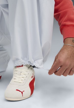 Vita elastiska byxor med en strukturerad yta är kombinerade med beiga och röda Puma-sneakers. En hand justerar byxans mudd.