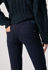 Mörk marinblå skinny jeans med två bakfickor och en märkesetikett. Tyget ser slätt ut med en lätt stretch. Matchas med en strukturstickad tröja.