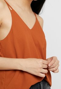 Camisole couleur rouille avec un décolleté en V, des fines bretelles et une texture douce. Le tissu est fluide, associé à un short en denim foncé.