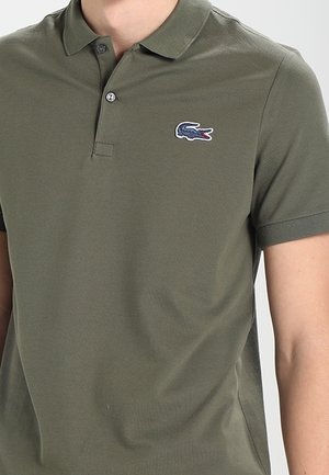 Polo verde oliva hecho de tela texturizada, con una tapeta de tres botones y un logo de cocodrilo bordado en el pecho.