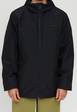 Uomo che indossa una giacca nera con cappuccio e zip frontale, con un logo discreto sul petto, abbinata a pantaloni verde oliva.